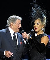 A man and a woman standing closely together. The man (left) is wearing a grey suit, white shirt and a black tie while the woman (right) is wearing a black gown, black gloves and a black headpiece. They both hold a microphone in their left hand.