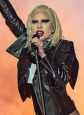 A blonde woman with wet-look hairstyle singing to a microphone on stage. She is wearing a black leather jacket.