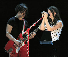 The White Stripes standing on stage: Meg is to the right, wearing a white polka dot shirt and black pants, singing into a mic; to her right is Jack, wearing a black shirt and red pants.