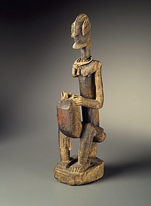 A wooden sculpture of a man seated on a low stool, playing a kora which rests on and slightly between his knees. The figure is stylized, with an elaborate hairstyle, the kora body slightly boxy.