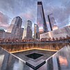 Illuminated water falls into the 9/11 Memorial south pool at sunset, and glass-clad One World Trade Center with other skyscrapers in the background