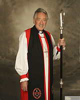 An Anglican bishop with a crosier, wearing a rochet under a red chimere and cuffs, a black tippet, and a pectoral cross
