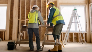 A male construction worker with a physical disability discusses blueprints with a female colleague over a worktable inside a home under construction.
